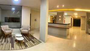 Een moderne lobby met een receptiebalie, een zithoek met stoelen en een kleine tafel, en een groot raam.