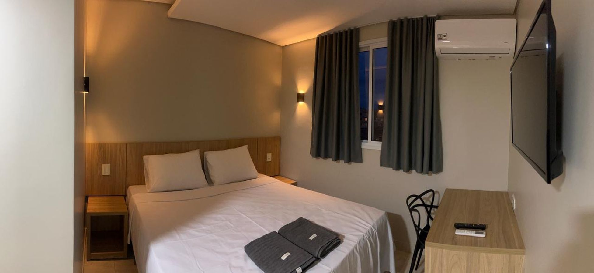 A hotel room with a bed, bedside tables, a desk with a chair, an air conditioning unit, and a TV mounted on the wall.