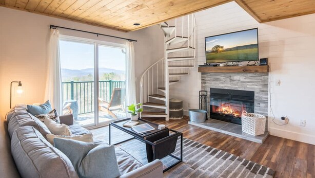 A modern living room with a fireplace, a large window, and a spiral staircase. - Mountain Views Indoor Pool Access Full Kitchen (Gatlinburg)