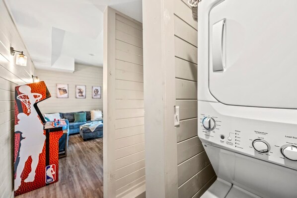 A laundry room with a white washing machine, a small table with a basketball-themed decoration, and a couch in the background. - NEW Lake Cabin Sleeps 14 Indoor Pool Hot Tub (Sevierville)