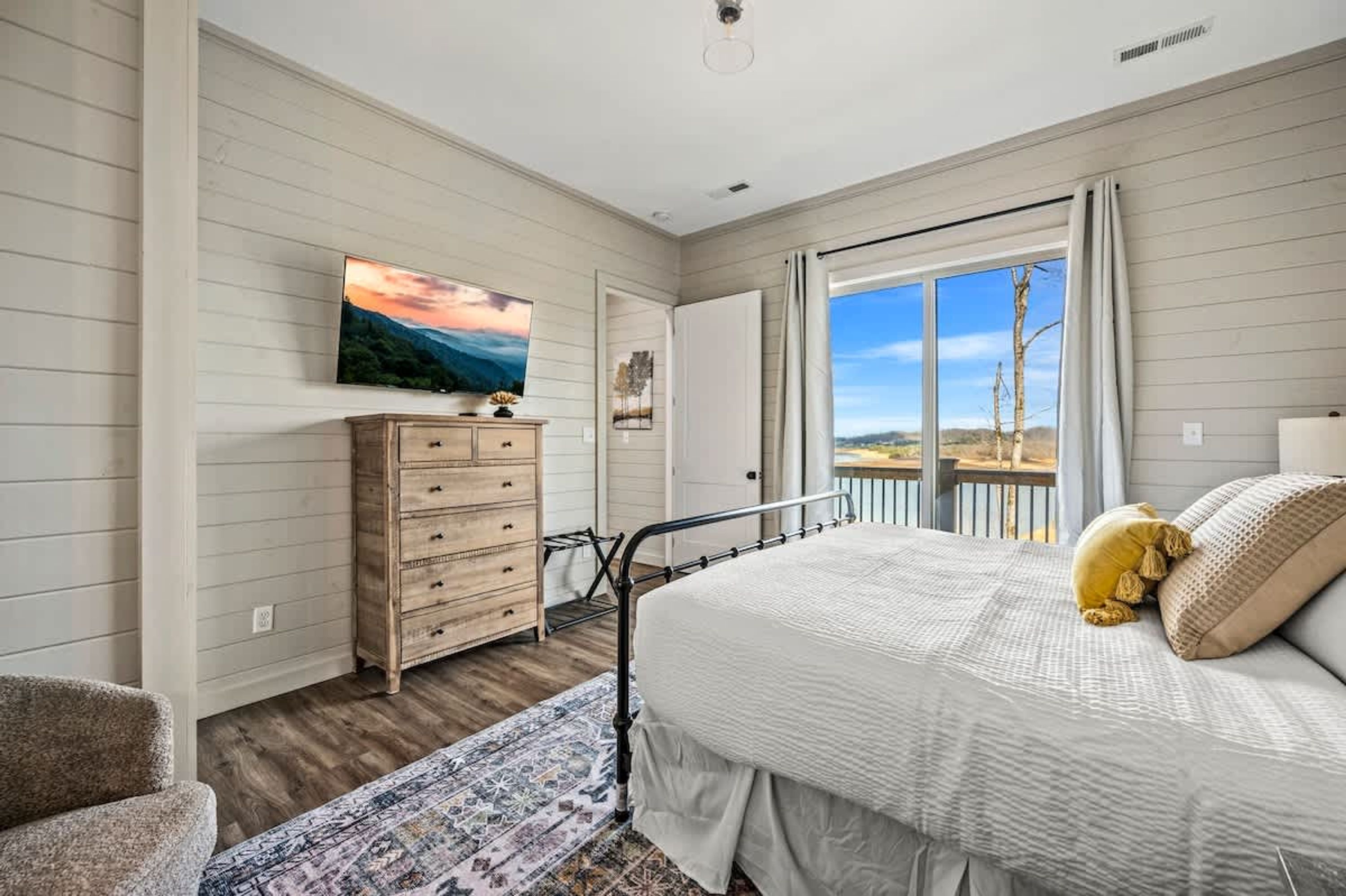Ein Schlafzimmer mit Bett, Kommode, Wandtelevison und Fenster mit Aussicht ins Freie.