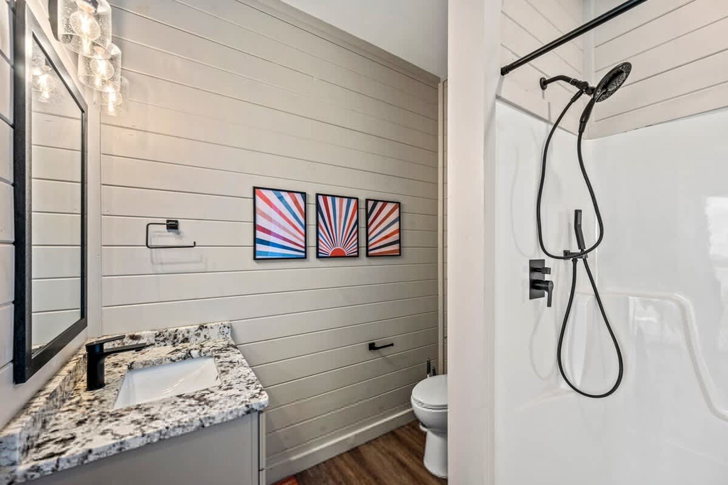 Une salle de bain avec un lavabo blanc, des toilettes et une douche. Trois œuvres d’art sont accrochées au mur au-dessus du lavabo.