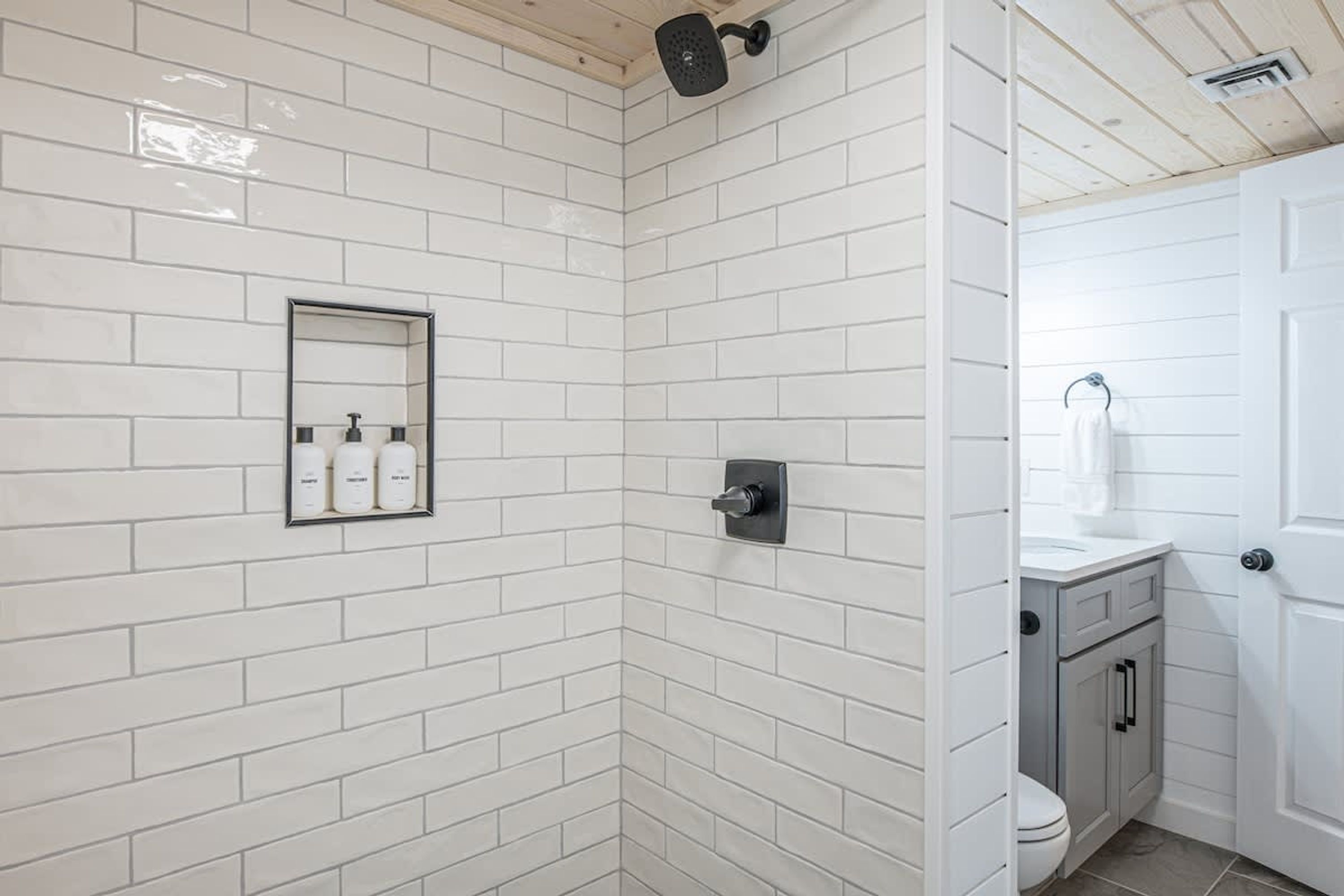 Une salle de bain avec des murs carrelés de blanc, une douche, des toilettes et un lavabo avec des meubles de rangement.