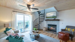A spacious living room with a fireplace, a ceiling fan, a staircase, and a large window.