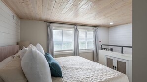 A bedroom with a wooden ceiling, a bed with white and blue bedding, two windows with blinds, and a small balcony.