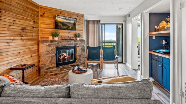 A cozy living room with a stone fireplace, wooden panel walls, and a view to the outside. - Two Bed Room Condo Mountain Views Indoor Pool (Gatlinburg)