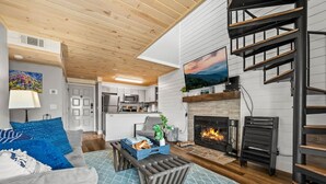A modern living room with a fireplace, a staircase, and a kitchen in the background. - Loft Sleeps 8 Views Indoor Pool Access (Gatlinburg)