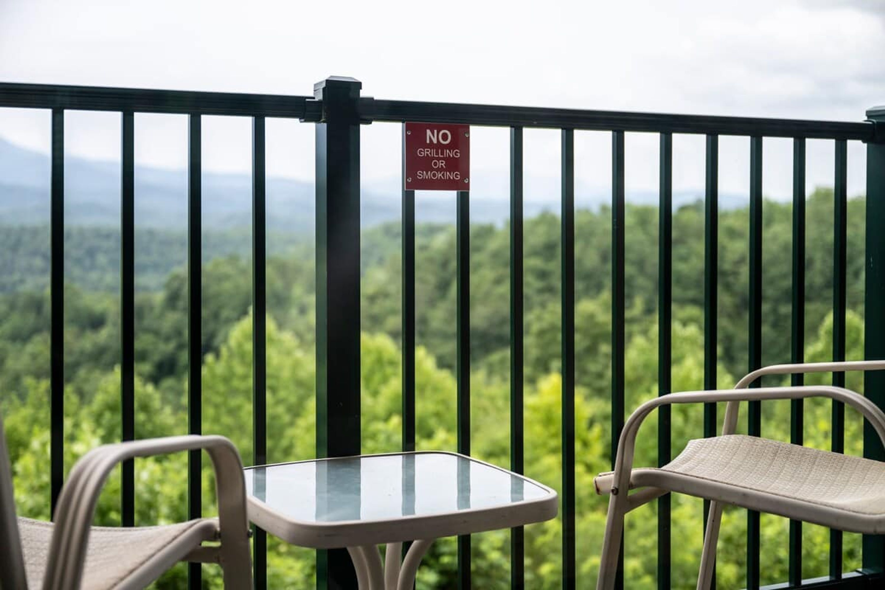A balcony with a 'NO GRILLING OR SMOKING' sign, a glass table, and two chairs, overlooking a forested landscape.