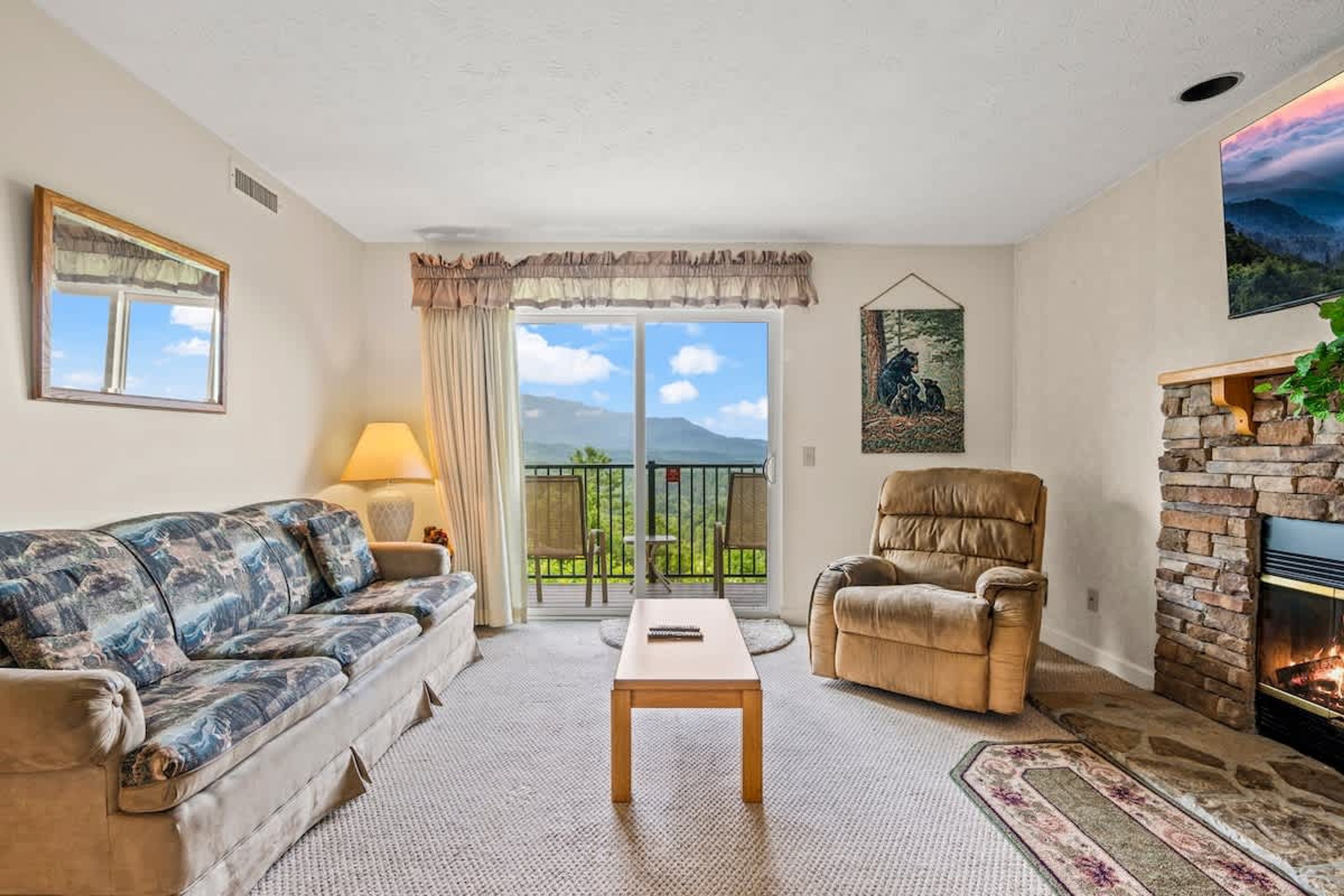 A living room with a sofa, armchair, coffee table, fireplace, and a balcony with a view of mountains.