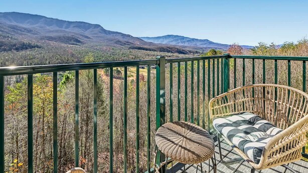 Un balcón con una silla de mimbre y una mesita, con vistas a un paisaje montañoso.