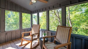 A porch with two rocking chairs, a ceiling fan, and large windows with a view of trees.