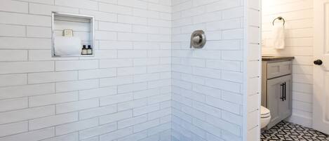 A bathroom with white subway tiles, a shower with a rain showerhead, a toilet with a white toilet paper holder, and a small shelf with toiletries.