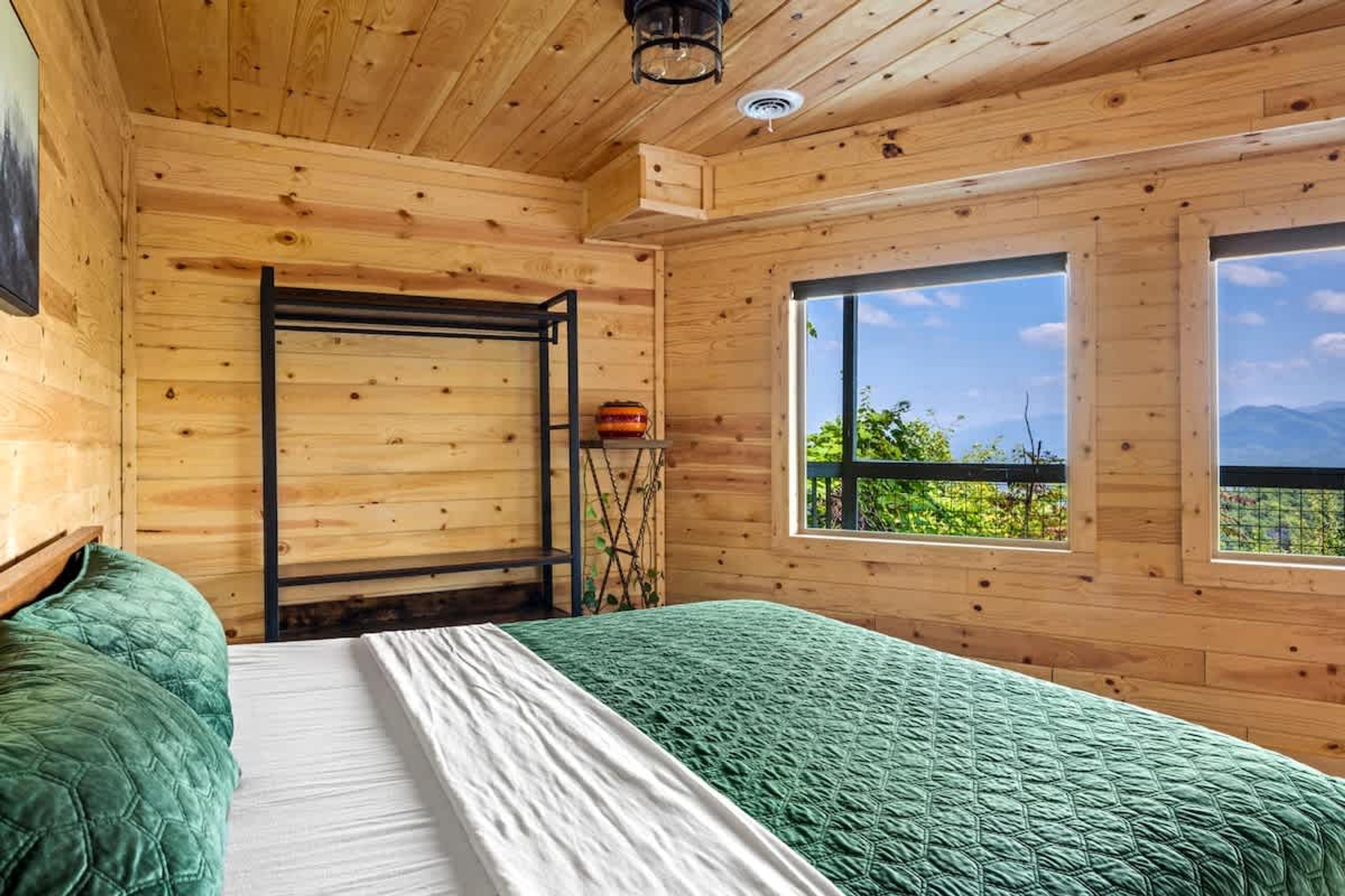 A wooden cabin bedroom with a bed, a window with a view of greenery and mountains, and a wooden ceiling.