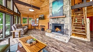 A cozy cabin interior with a stone fireplace, wooden floors, and a large window overlooking greenery. - 180 Views Sleeps 7 Private Hot Tub Full Kitchen (Gatlinburg)