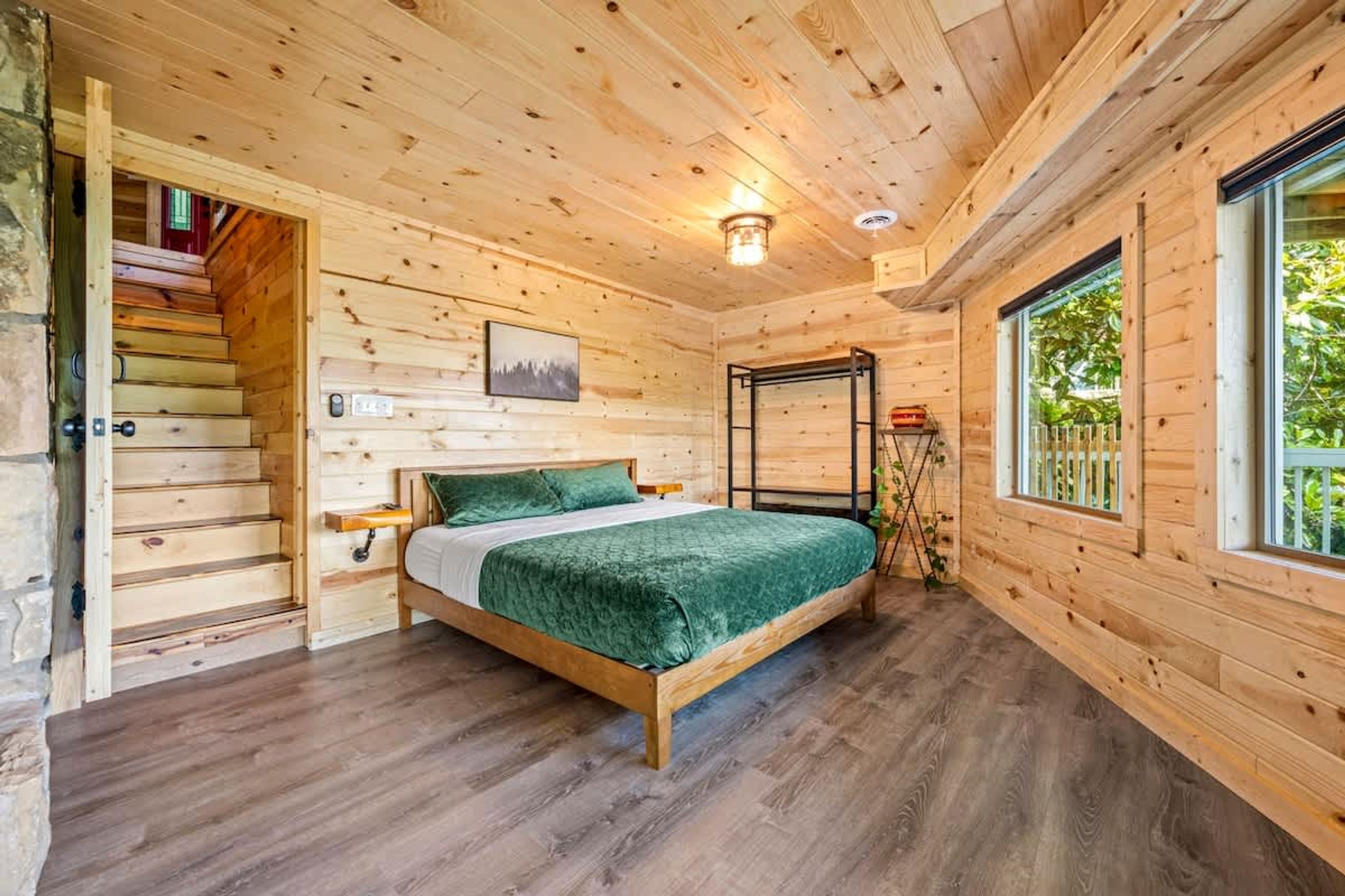 A wooden cabin bedroom with a bed, a staircase, a window, and a wall-mounted light fixture.