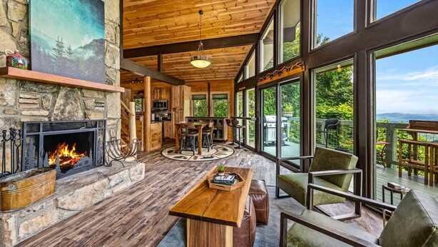 A spacious living area with a fireplace, wooden furniture, and large windows offering a view of the outdoors. - 180 Views Sleeps 7 Private Hot Tub Full Kitchen (Gatlinburg)