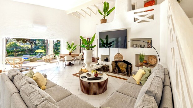 A modern living room with a large sofa, a round coffee table, and a TV mounted on the wall. There are potted plants and a fireplace. - Spectacular Villa in Quinta do Lago - Private Pool, BBQ & 5-Star Concierge (Faro)