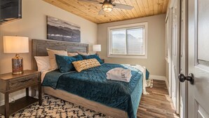 A bedroom with a wooden ceiling, a bed with a blue quilt, a painting, a window with blinds, and a door. - Indoor Pool Access Full Kitchen Mountain Views (Gatlinburg)