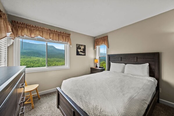 A bedroom with a large bed, a desk, a chair, a nightstand with a lamp, and a window with curtains.