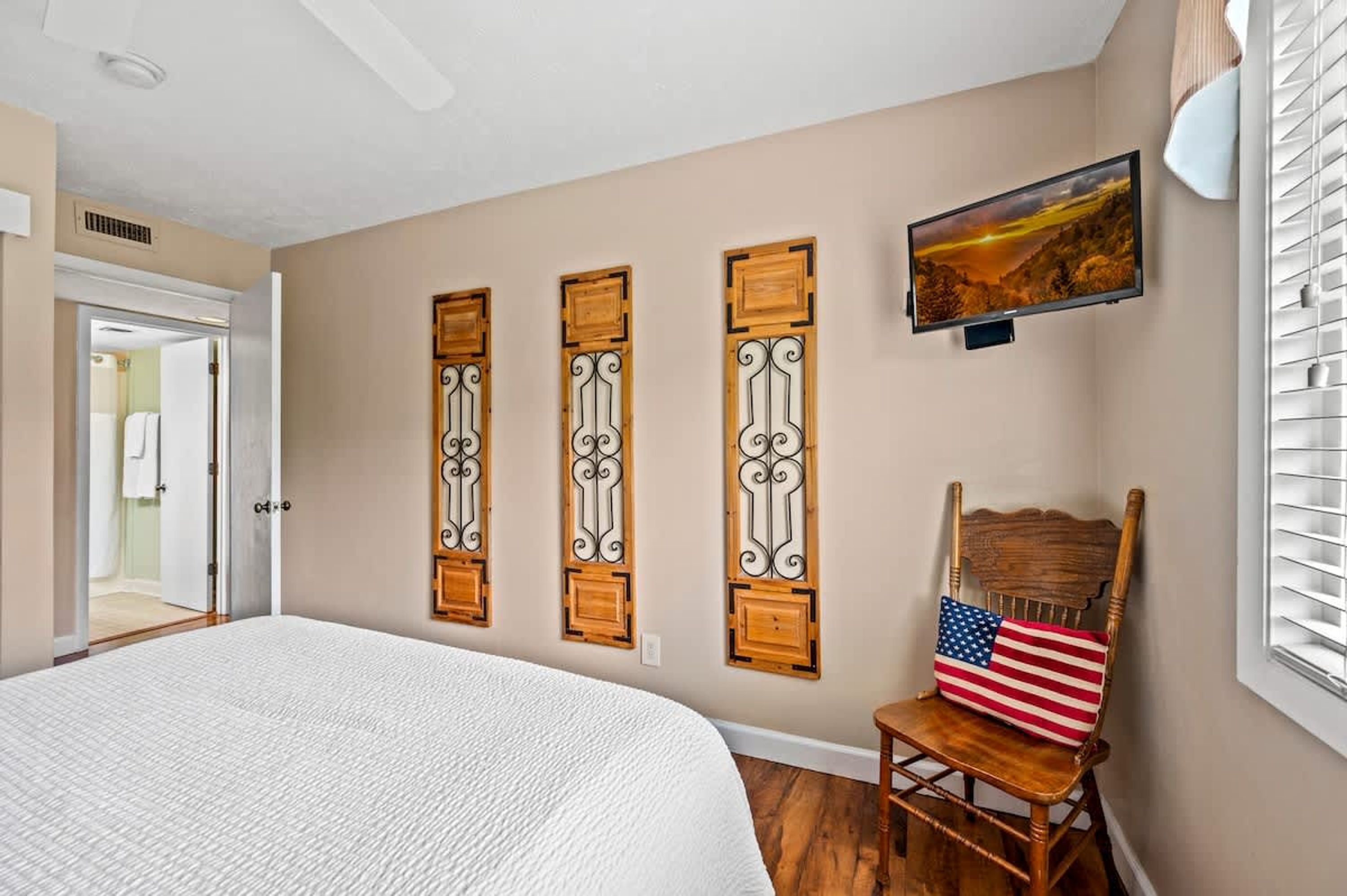 Un dormitorio con cama, televisor montado en la pared y una silla con almohada de la bandera estadounidense.