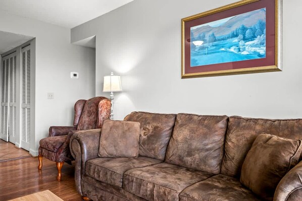 A living room with a brown leather sofa, a chair, and a painting on the wall. - Indoor Pool Hot Tub Sauna Sleeps 6 Full Kitchen (Gatlinburg)