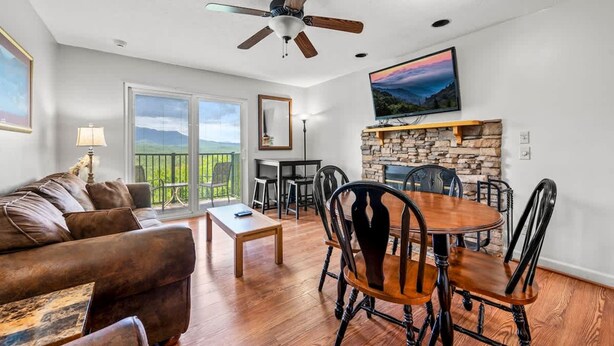 A spacious living room with a fireplace, a dining area with wooden chairs, a ceiling fan, and a large window with a view. - Indoor Pool Hot Tub Sauna Sleeps 6 Full Kitchen (Gatlinburg)