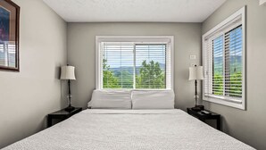 A bedroom with a large bed, two bedside tables with lamps, a window with blinds, and a view of greenery outside. - Private Balcony w Views Heated Pool Full Kitchen (Gatlinburg)