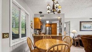 A dining area with a wooden table and chairs, a kitchen in the background, and a window with blinds. - Private Balcony w Views Heated Pool Full Kitchen (Gatlinburg)