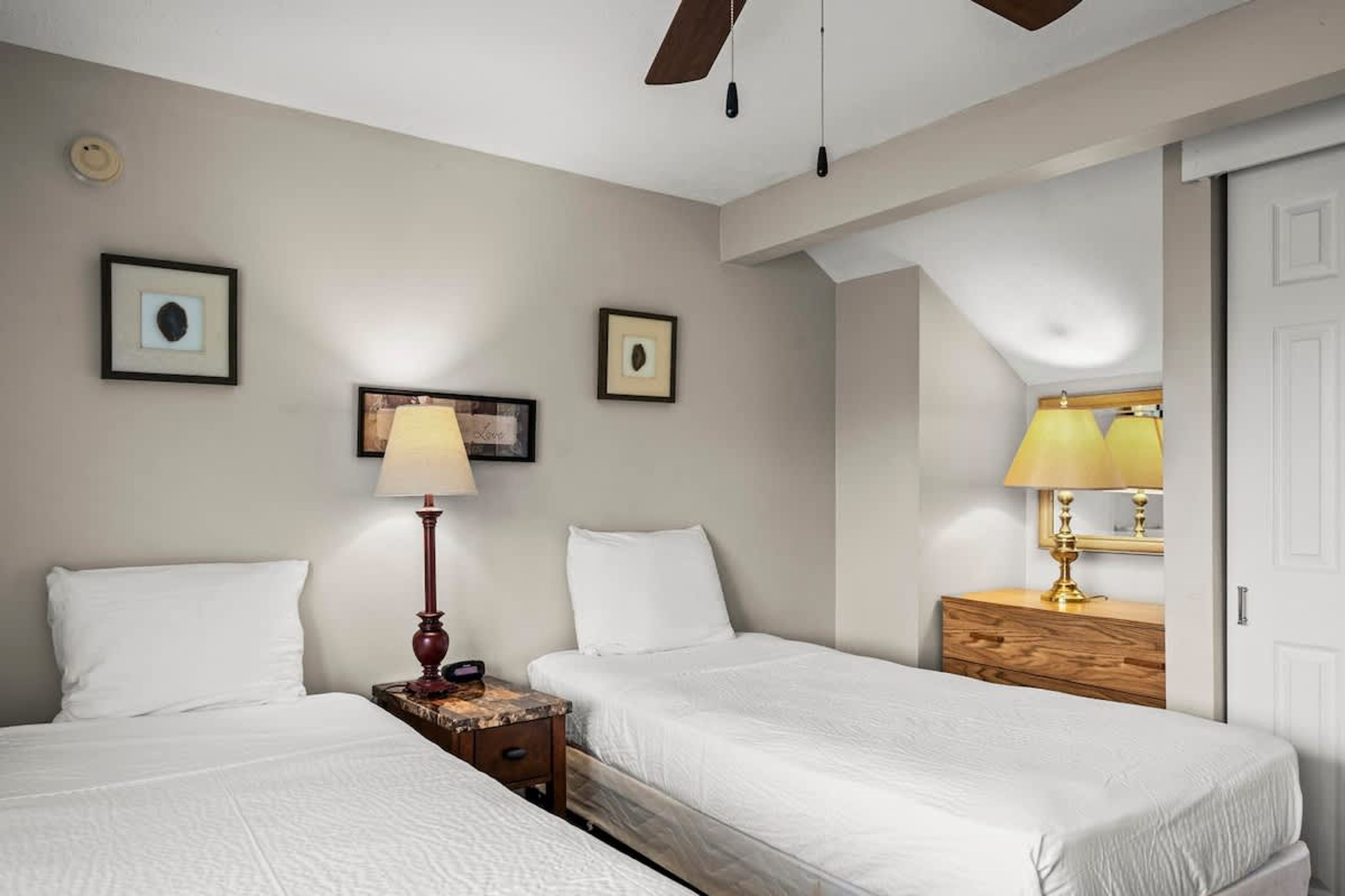 Habitación de hotel con dos camas, una mesita de noche de madera con lámpara y un aparador con espejo.