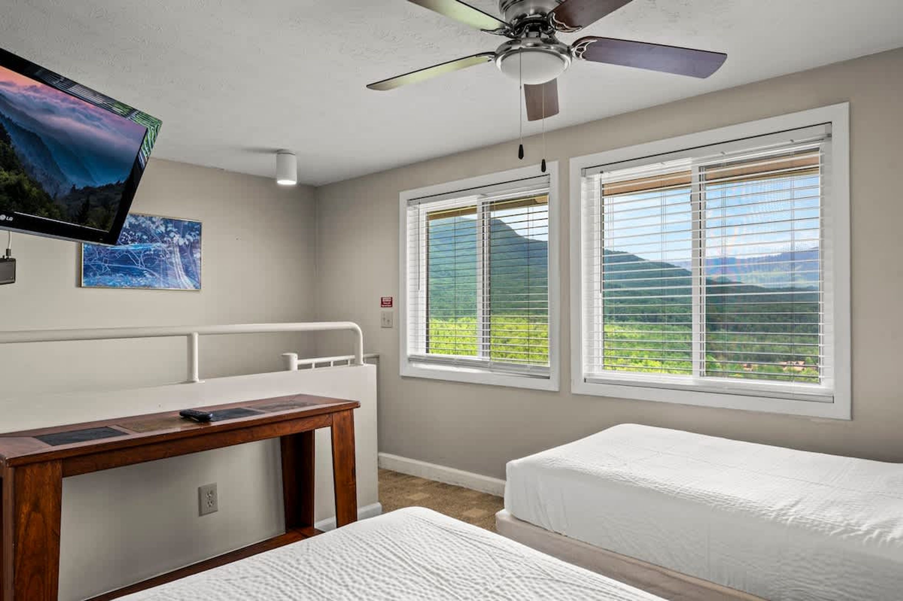A bedroom with two beds, a desk, a ceiling fan, and large windows with a view of greenery.