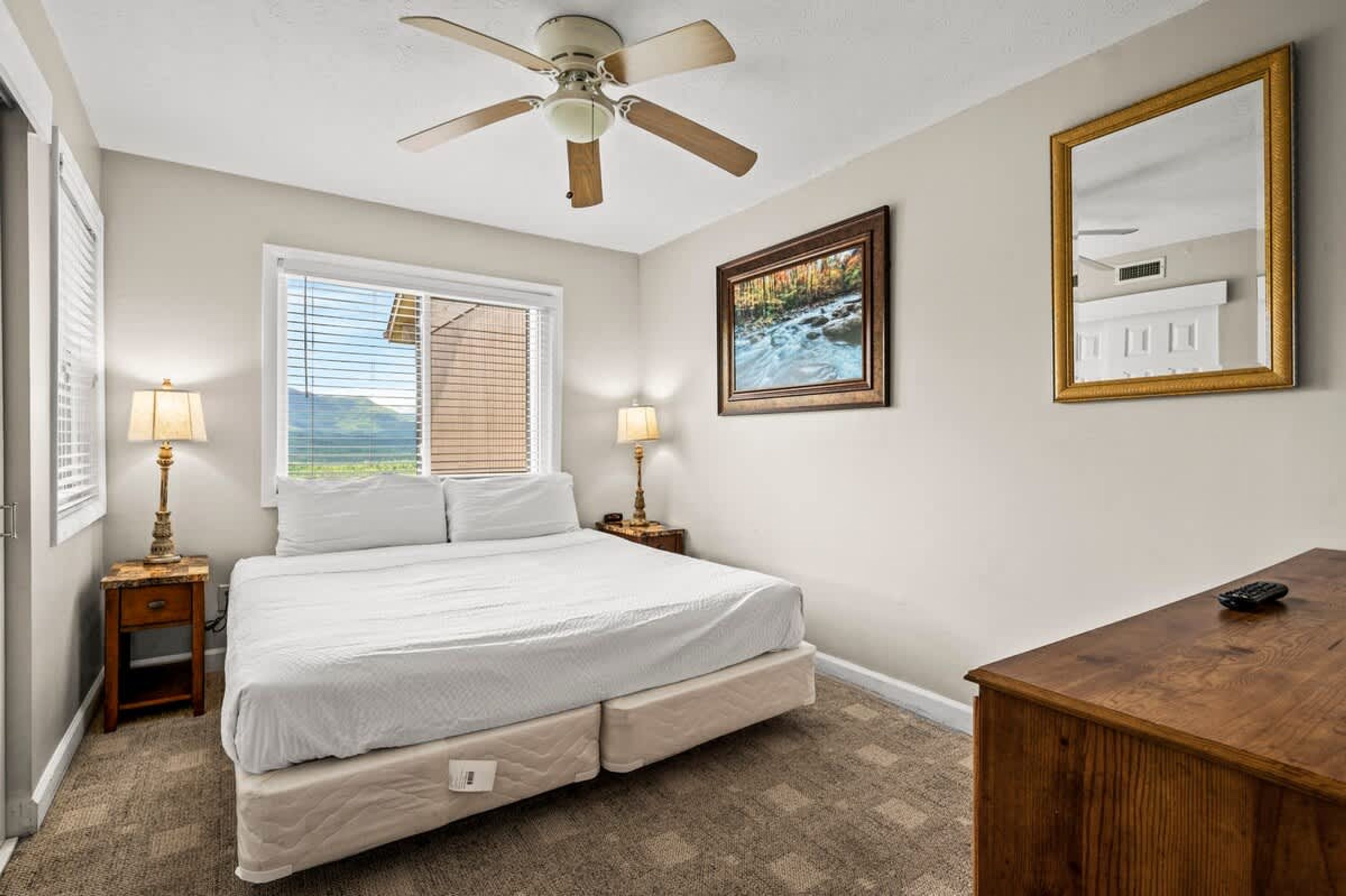 A bedroom with a bed, bedside lamps, a ceiling fan, a window with blinds, a painting, a mirror, and a wooden desk.