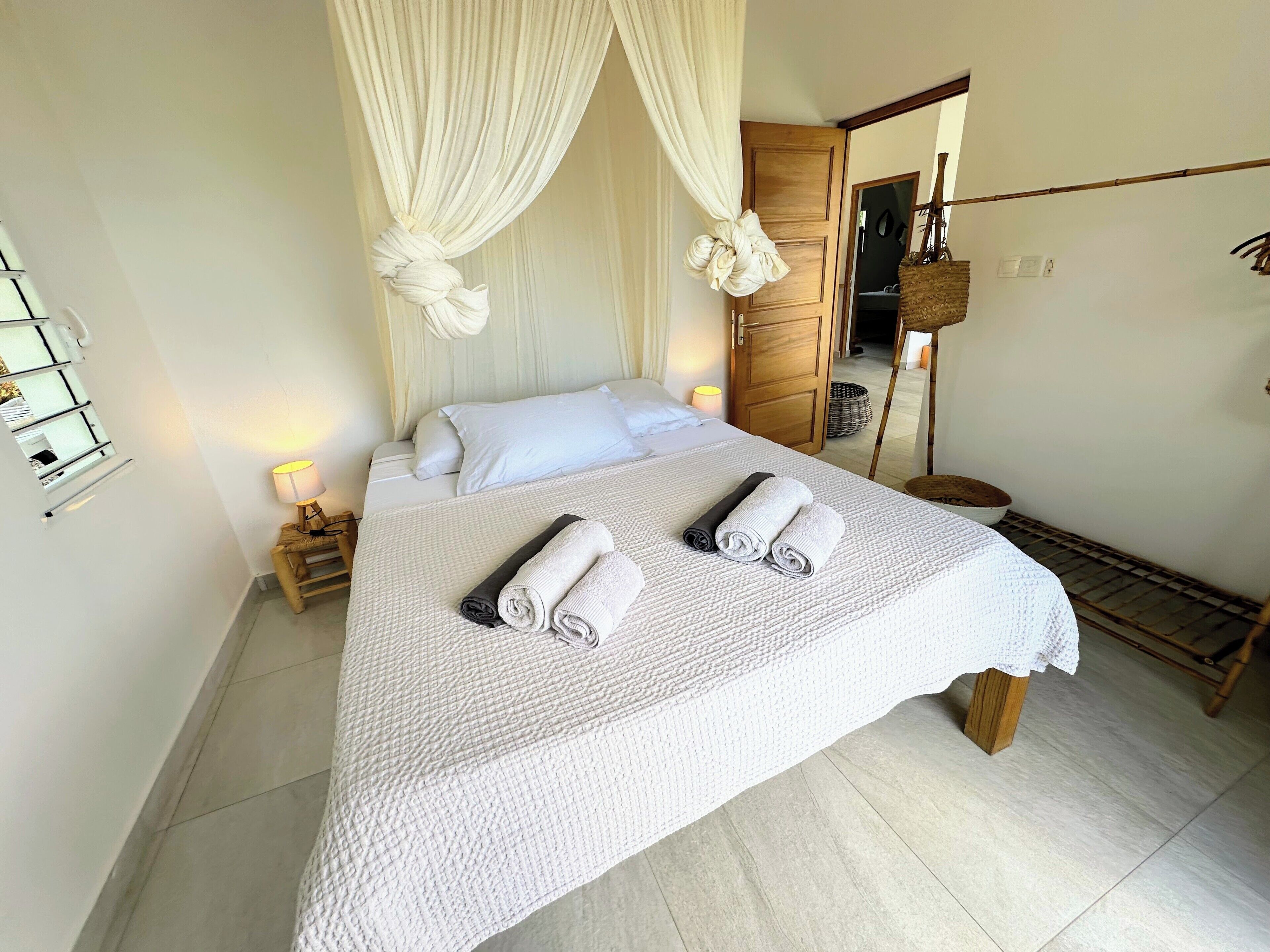 A neatly made bed with a white bedspread and two rolled towels, a wooden wardrobe, a bedside lamp, and a mirror on the wall.