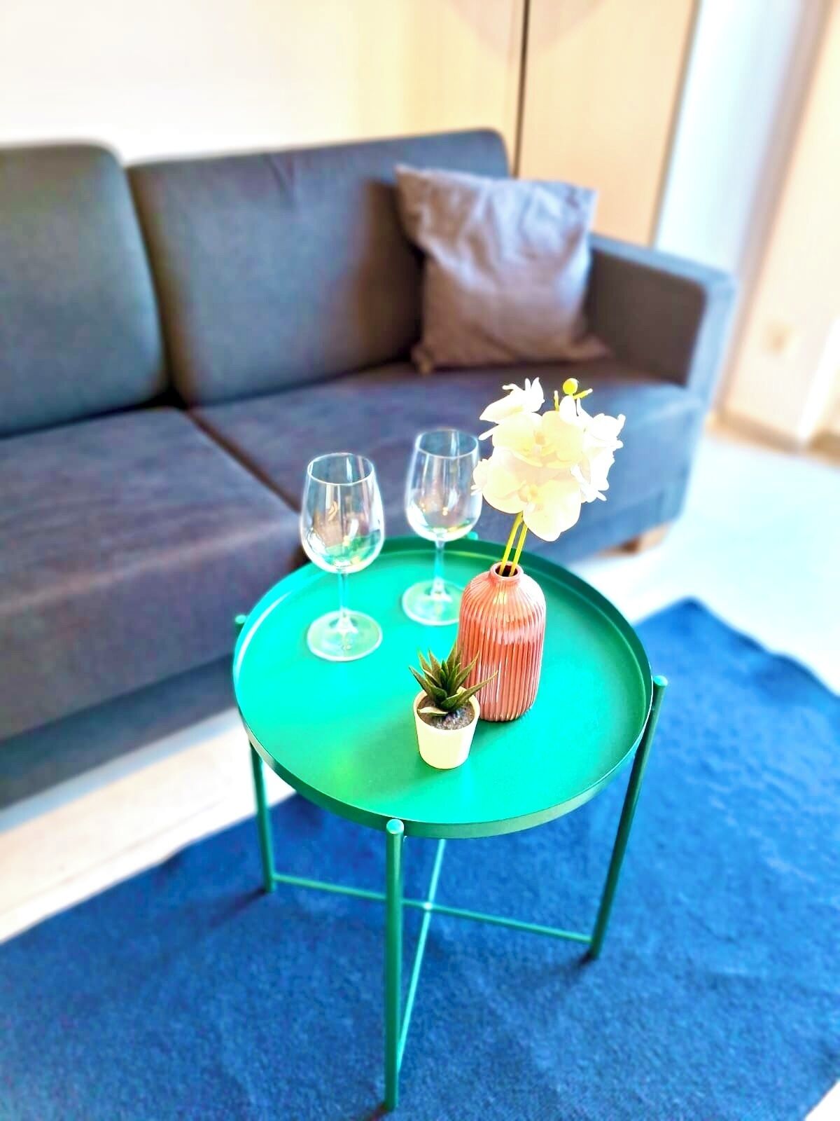 Una mesa de centro redonda verde con tres copas de vino y un florero con flores blancas, ubicada frente a un sofá gris con un cojín marrón.