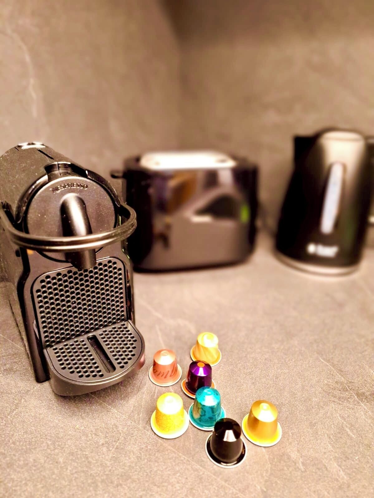 Una cafetera con un recipiente para goteo y varias cápsulas de café de colores sobre una superficie texturizada.