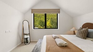 Een slaapkamer met een groot raam, een bed met een beige deken, een nachtkastje met een lamp en een spiegel in volle lengte.