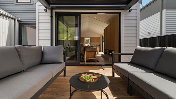 Een modern buitenzitgedeelte met een houten tafel en een bank, een glazen deur naar een binnenruimte, en een houten terras.