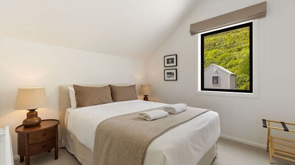 A bedroom with a bed, two bedside tables with lamps, a window with a view of greenery, and a wooden bench.