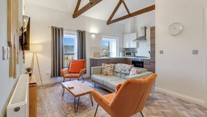 A modern living room with a sofa, two armchairs, a coffee table, and a floor lamp. There is a kitchen in the background with a microwave, stove, and cabinets. - Chai- Tea Warehouse - North East Escapes (Alnwick)
