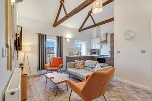 Una sala de estar moderna con un sofá, dos sillones, una mesa de centro y una lámpara de pie. Se observa una cocina al fondo con microondas, estufa y armarios.