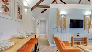 A modern dining area with a long wooden table set for a meal, orange cushioned bar stools, and a television mounted on the wall. - Chai- Tea Warehouse - North East Escapes (Alnwick)