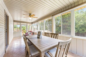Une terrasse couverte avec une table et des chaises en bois, de grandes fenêtres et un ventilateur de plafond.