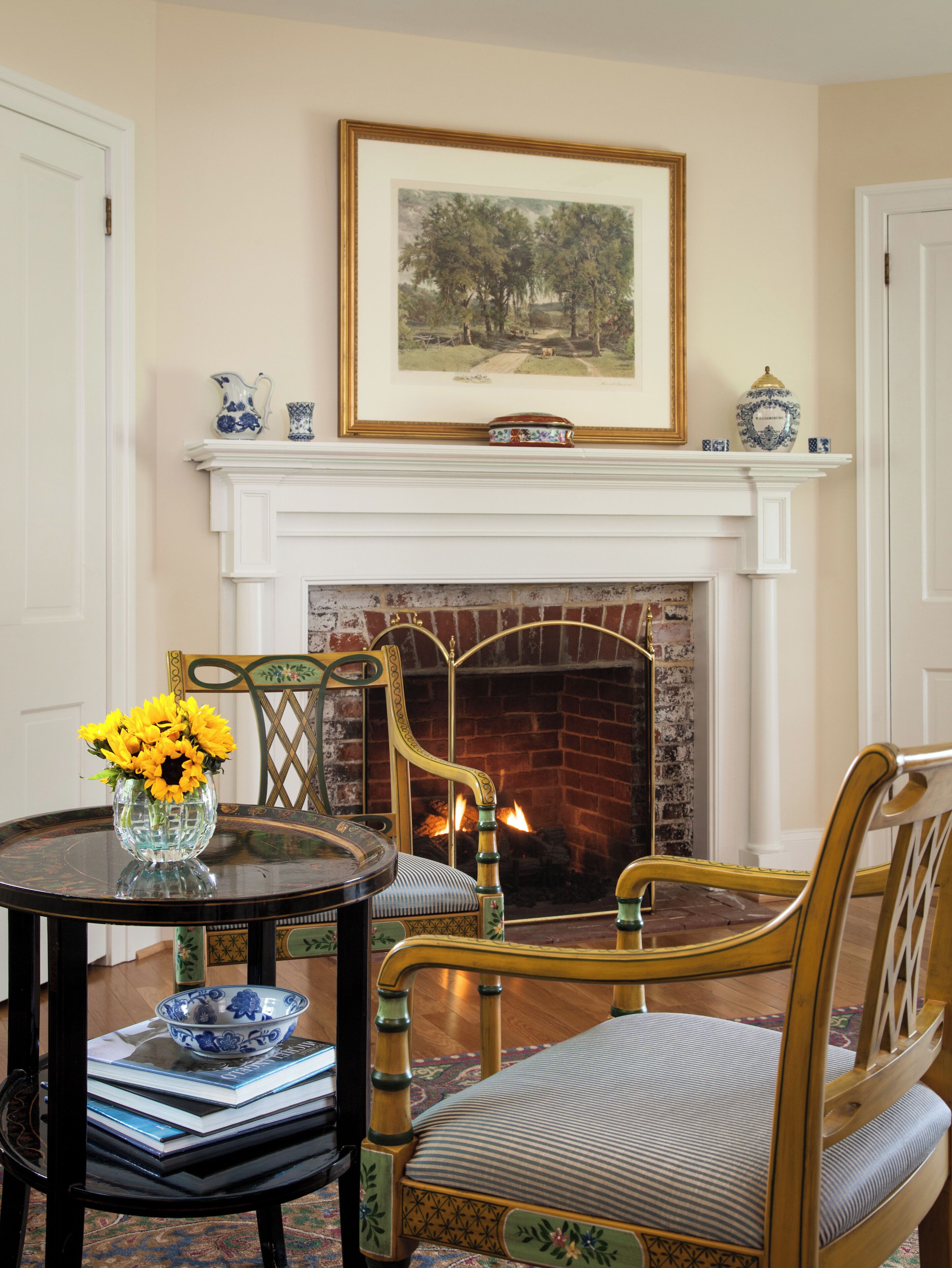 A room with a fireplace, a framed painting above it, and a table with books and a vase of flowers.