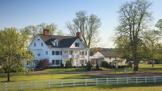 Une grande maison blanche avec un toit sombre, de nombreuses cheminées et une clôture blanche au premier plan.