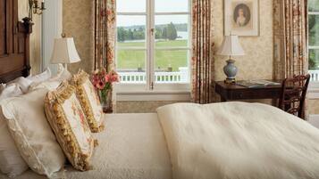 A bedroom with a large bed, two bedside lamps, a desk with a chair, a vase of flowers, and a view of a lake and white fence.