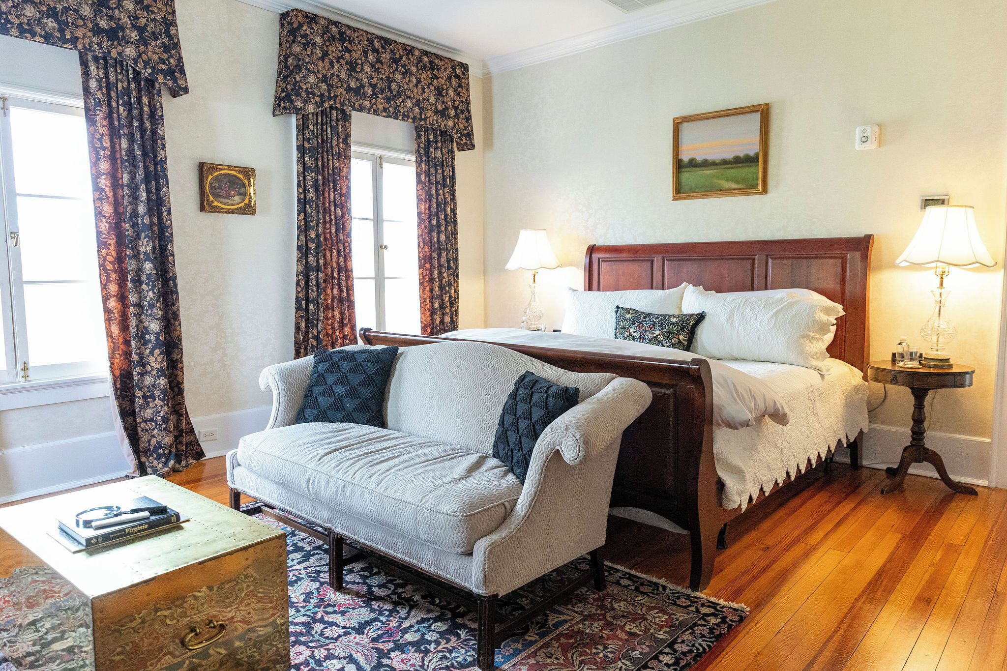 A hotel room with a wooden bed, two bedside lamps, a floral-patterned sofa, a small table with a book, and a patterned rug.