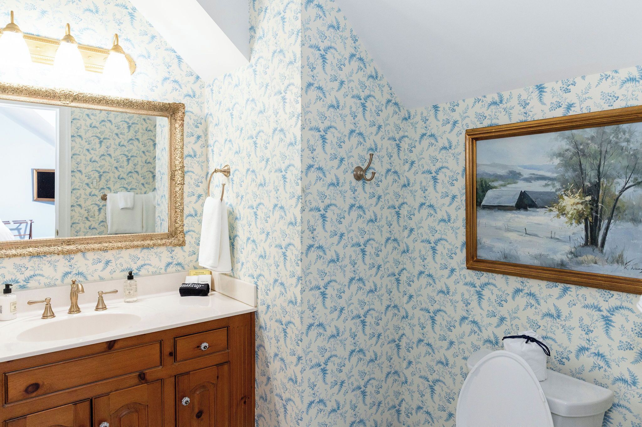 A bathroom with floral wallpaper, a large mirror, a sink with toiletries, and a painting on the wall.