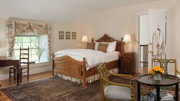 Une chambre à coucher de style traditionnel, avec un lit en bois, deux tables de chevet avec des lampes, une chaise, une petite table ronde ornée d’un vase, et une vue sur l’extérieur par une fenêtre avec des rideaux.