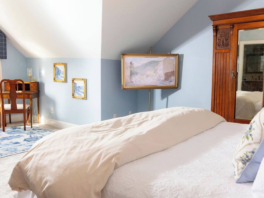 A bedroom with a bed, a wooden wardrobe, a desk with a chair, and framed artwork on the wall.