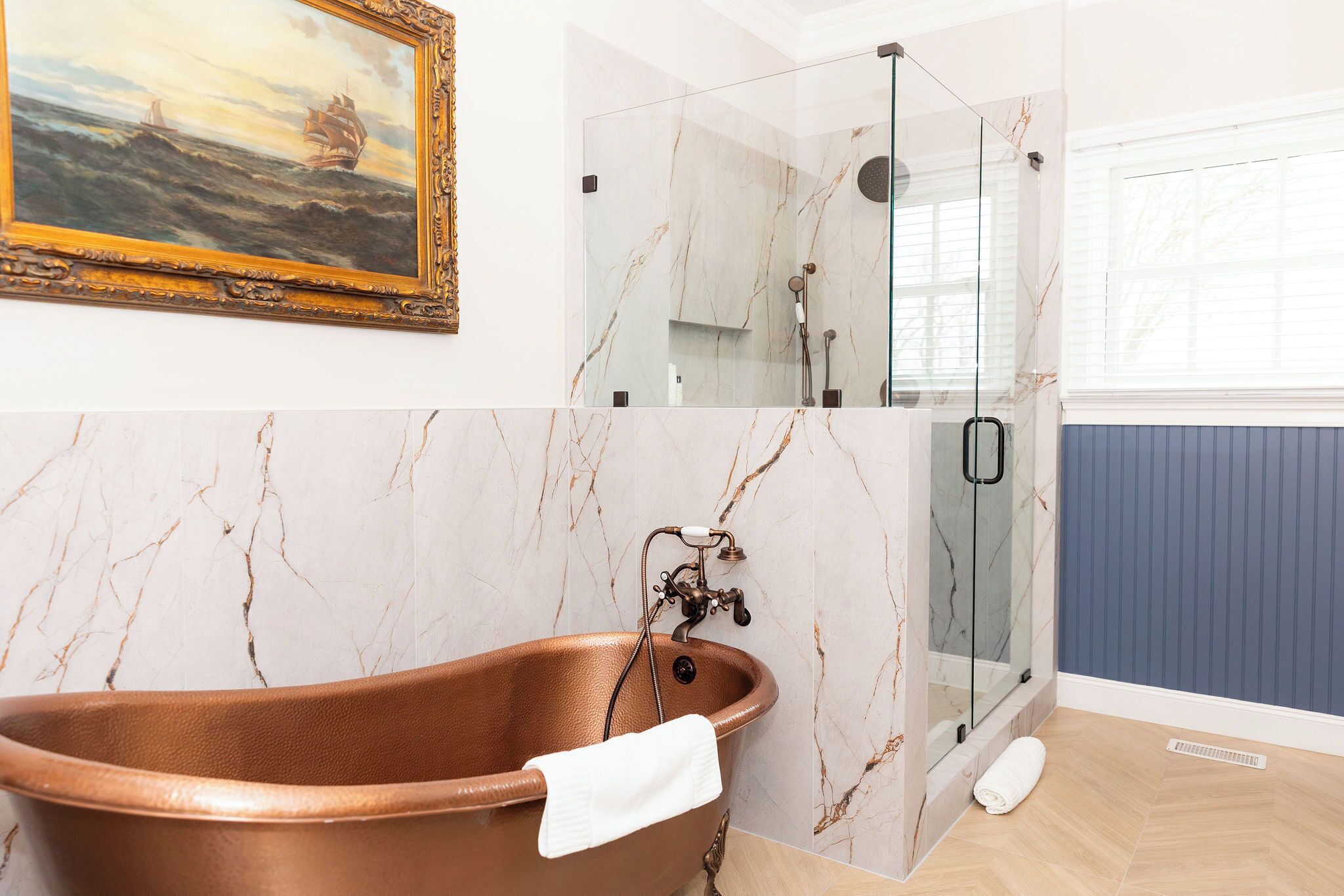 A bathroom with a freestanding copper bathtub, a glass-enclosed shower, and a marble wall with a framed seascape painting.