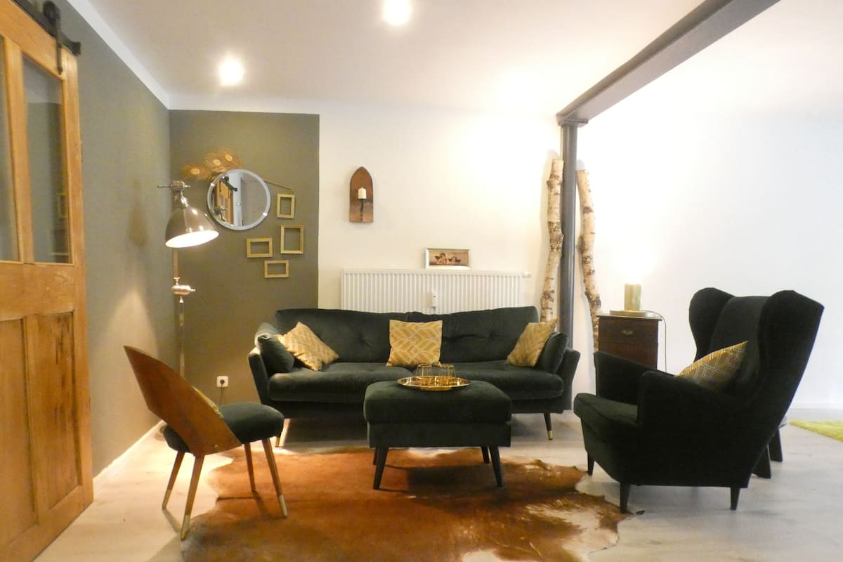 Ein Wohnzimmer mit einem dunkelgrünen Sofa, zwei Sesseln, einem Couchtisch, einem Holzsessel, einem Kuhfellteppich, einem Spiegel, gerahmten Bildern und einer Heizung.